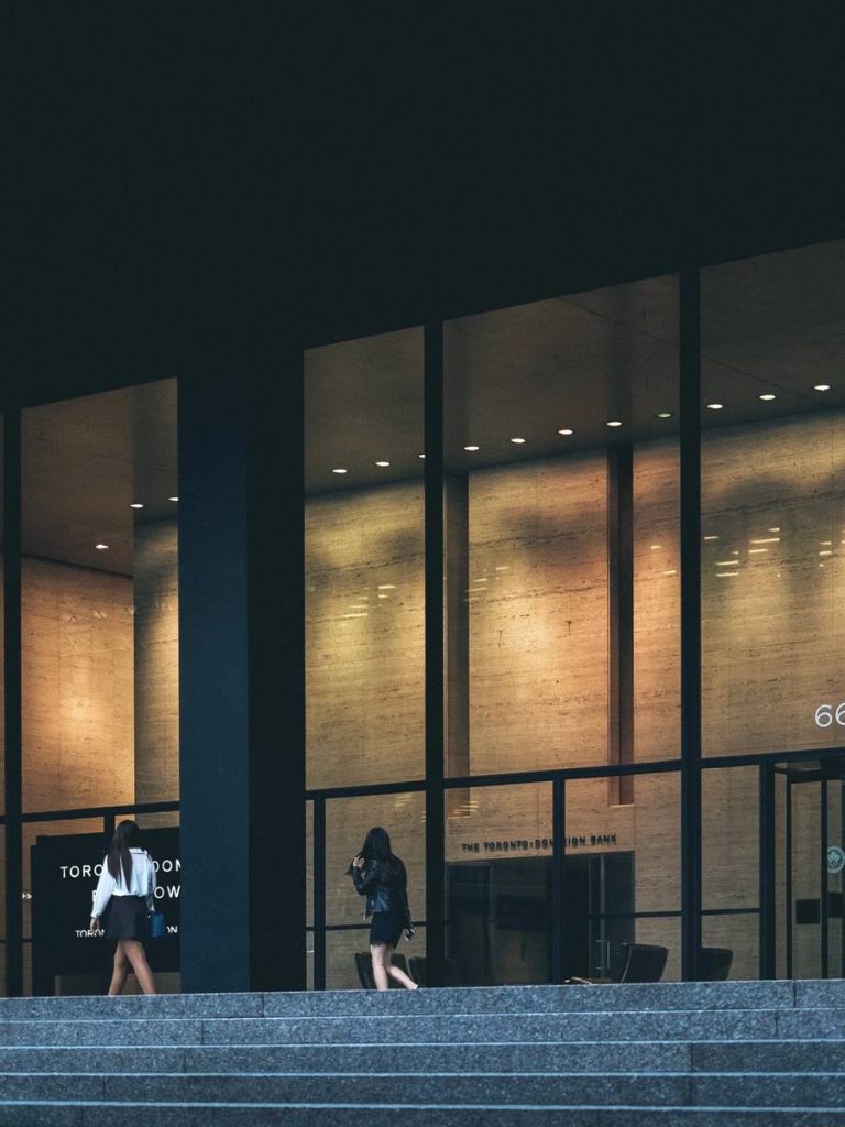 Two women walking past a modern building with large glass windows and warm interior lighting.